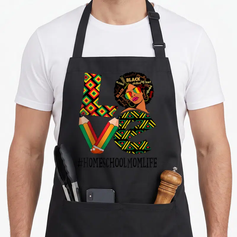 Homeschool african apron - Cartoon woman with word-filled afro, patterned LOVE letters, two crossed pencils — shown on a black cotton canvas apron against a white background