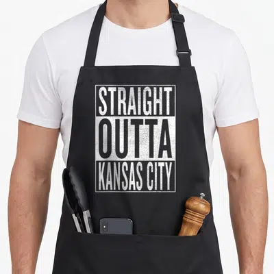 Kansas travel aprons - Stacked distressed text reading STRAIGHT OUTTA KANSAS CITY in white and grey blocks — shown on a black cotton canvas apron against a white background