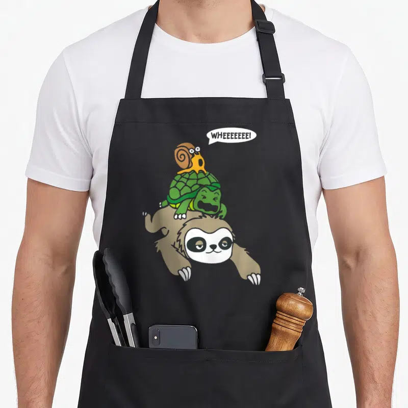 Turtle sloth apron - Cartoon brown sloth carrying green turtle and orange snail with speech bubble text WHEEEEEEEI — shown on a black cotton canvas apron against a white background
