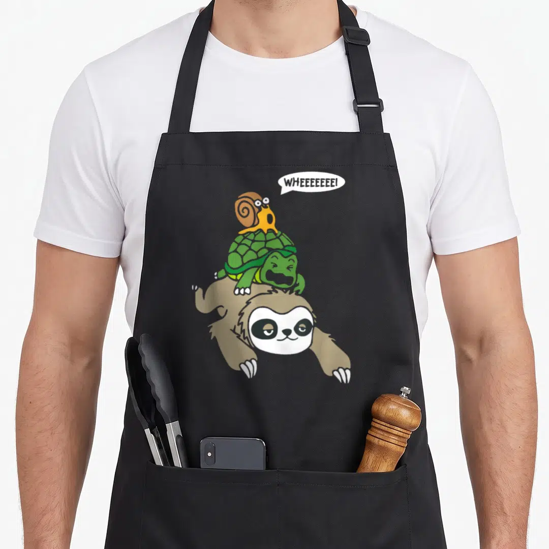 Turtle sloth apron - Cartoon brown sloth carrying green turtle and orange snail with speech bubble text WHEEEEEEEI — shown on a black cotton canvas apron against a white background