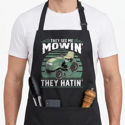 Riding see me aprons - Distressed green riding mower with THEY SEE ME MOWIN THEY HATIN text over stripes — shown on a black cotton canvas apron against a white background