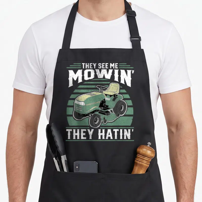Riding see me aprons - Distressed green riding mower with THEY SEE ME MOWIN THEY HATIN text over stripes — shown on a black cotton canvas apron against a white background