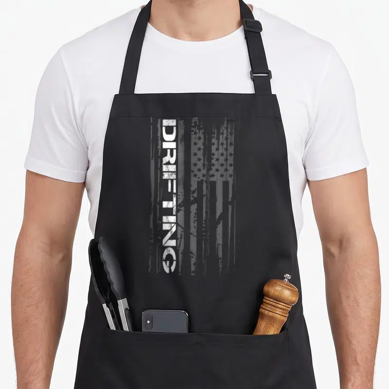 Car racing american flag apron - Vertical DRIFTING text over distressed white and grey USA flag graphic — shown on a black cotton canvas apron against a white background