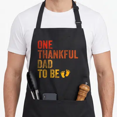 Thankful pregnancy announcement thanksgiving apron - Stacked red orange and yellow text reading ONE THANKFUL DAD TO BE — shown on a black cotton canvas apron against a white background
