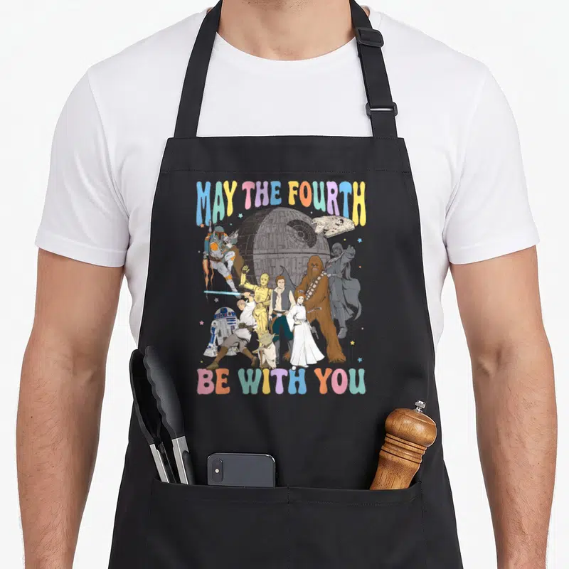 Funny may fourth apron - Retro Star Wars characters with MAY THE FOURTH BE WITH YOU text in blue pink yellow grey — shown on a black cotton canvas apron against a white background
