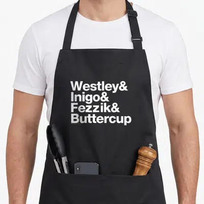 Westley princess apron - Stacked white text listing Westley, Inigo, Fezzik, and Buttercup with ampersands — shown on a black cotton canvas apron against a white background