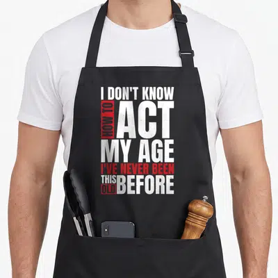 Vintage act age apron - Stacked white and red distressed text reading I don't know how to act my age I've never been — shown on a black cotton canvas apron against a white background