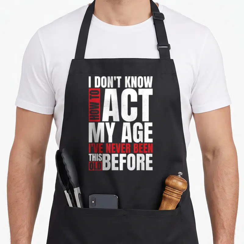 Vintage act age apron - Stacked white and red distressed text reading I don't know how to act my age I've never been — shown on a black cotton canvas apron against a white background