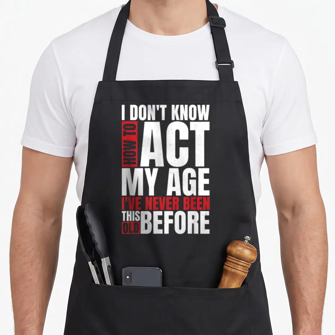 Vintage act age apron - Stacked white and red distressed text reading I don't know how to act my age I've never been — shown on a black cotton canvas apron against a white background