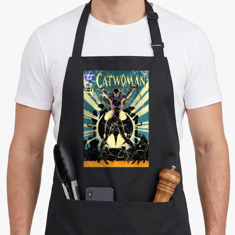 Cover catwoman apron - CATWOMAN text above standing figure in purple suit with black cats and yellow moon — shown on a black cotton canvas apron against a white background