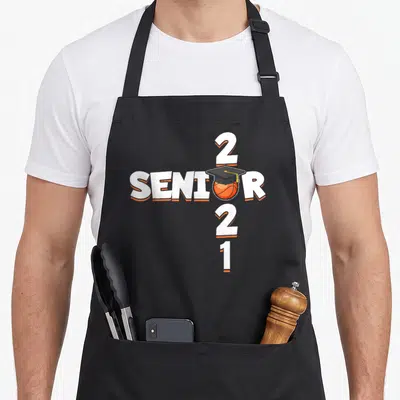Senior class player apron - White and orange 2021 text with basketball wearing a black graduation cap — shown on a black cotton canvas apron against a white background