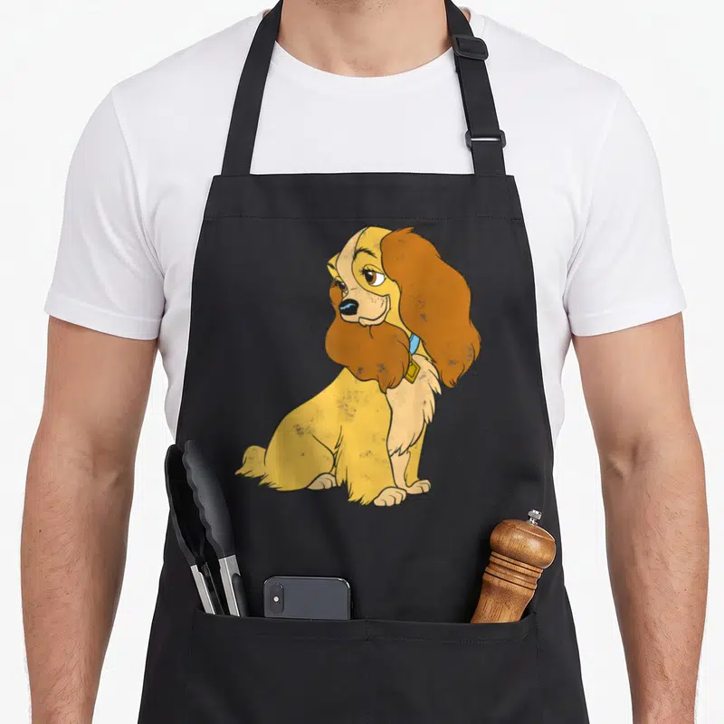 Simple lady and the tramp apron - Seated cartoon spaniel in yellow brown and blue with blue collar — shown on a black cotton canvas apron against a white background