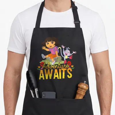 Adventure explorer apron - Dora and Boots sitting on grassy rock with Adventure AWAITS text in yellow and pink script — shown on a black cotton canvas apron against a white background