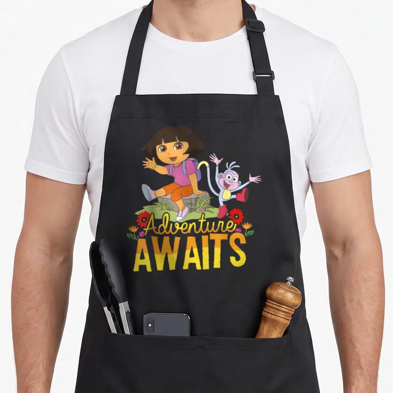 Adventure explorer apron - Dora and Boots sitting on grassy rock with Adventure AWAITS text in yellow and pink script — shown on a black cotton canvas apron against a white background