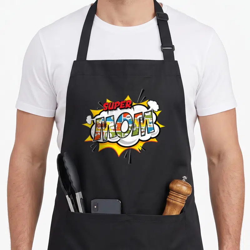 Comic mother's apron - SUPER MOM text in red above comic-panel filled letters inside a white cloud and yellow burst — shown on a black cotton canvas apron against a white background