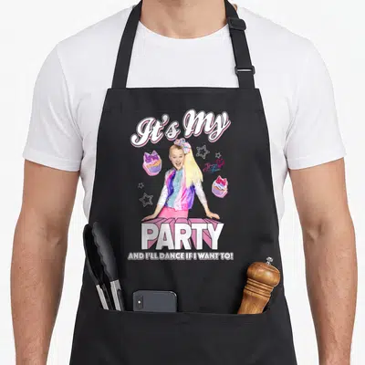 Funny occasion novelty apron - Girl centered between It's My Party text with cupcakes and stars in pink purple white — shown on a black cotton canvas apron against a white background