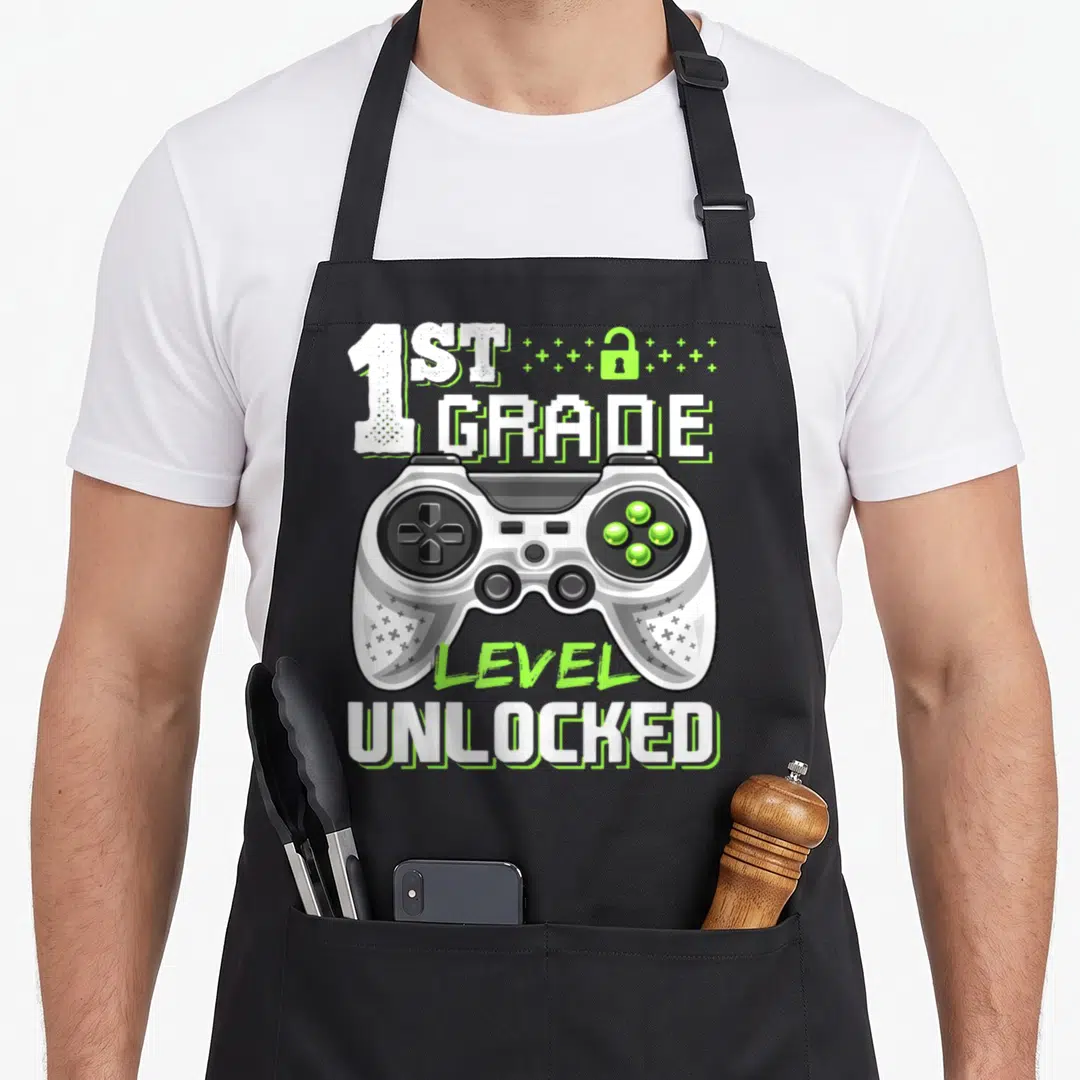 1st video game apron - White and lime green game controller with 1st Grade Level Unlocked text and lock symbol — shown on a black cotton canvas apron against a white background