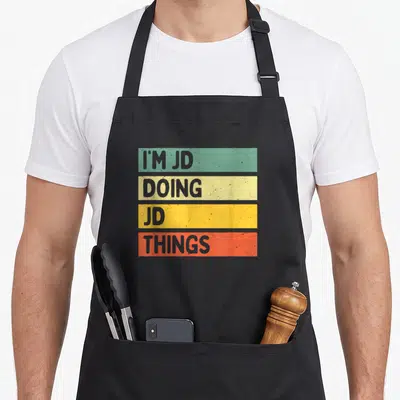 Doing jd apron - I'M JD DOING JD THINGS stacked text in green yellow orange and red retro blocks — shown on a black cotton canvas apron against a white background
