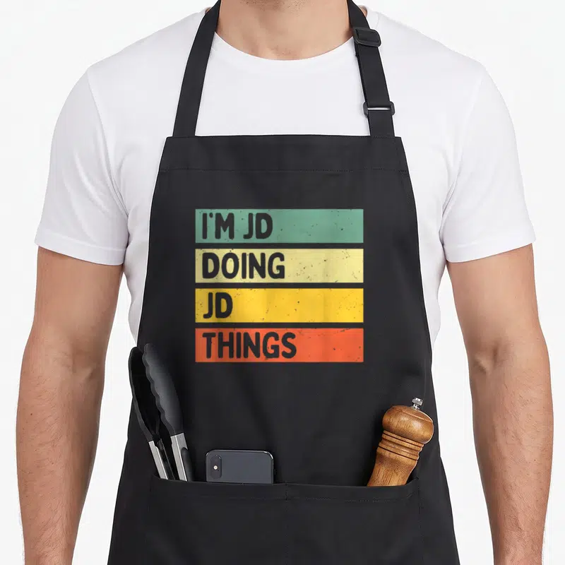 Doing jd apron - I'M JD DOING JD THINGS stacked text in green yellow orange and red retro blocks — shown on a black cotton canvas apron against a white background