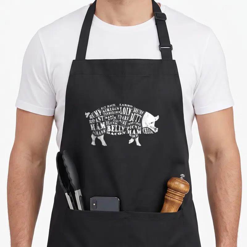 Pig barbecue apron - White distressed pig silhouette formed by text labels Rump Sirloin Lardo Loin Boston Butt — shown on a black cotton canvas apron against a white background