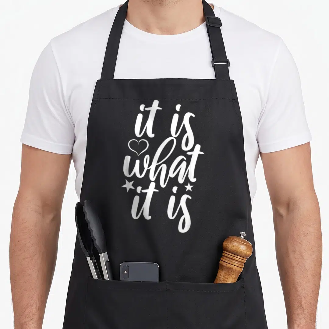 General gag apron - White script text it is what it is with heart and two stars — shown on a black cotton canvas apron against a white background