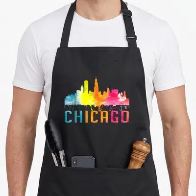 Watercolour chicago apron - Rainbow watercolor skyline above CHICAGO text in blue, yellow, orange, pink, and purple — shown on a black cotton canvas apron against a white background