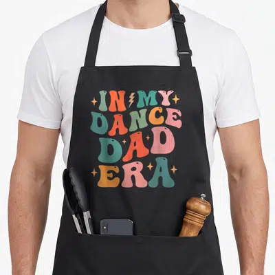 Era dance dad apron - Retro stacked text reading IN MY DANCE DAD ERA in teal, orange, pink, and green with a lightning — shown on a black cotton canvas apron against a white background