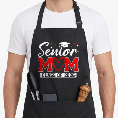 Senior proud graduate mom class apron - Red and white Senior MOM Class of 2026 text with heart and cap graphic — shown on a black cotton canvas apron against a white background