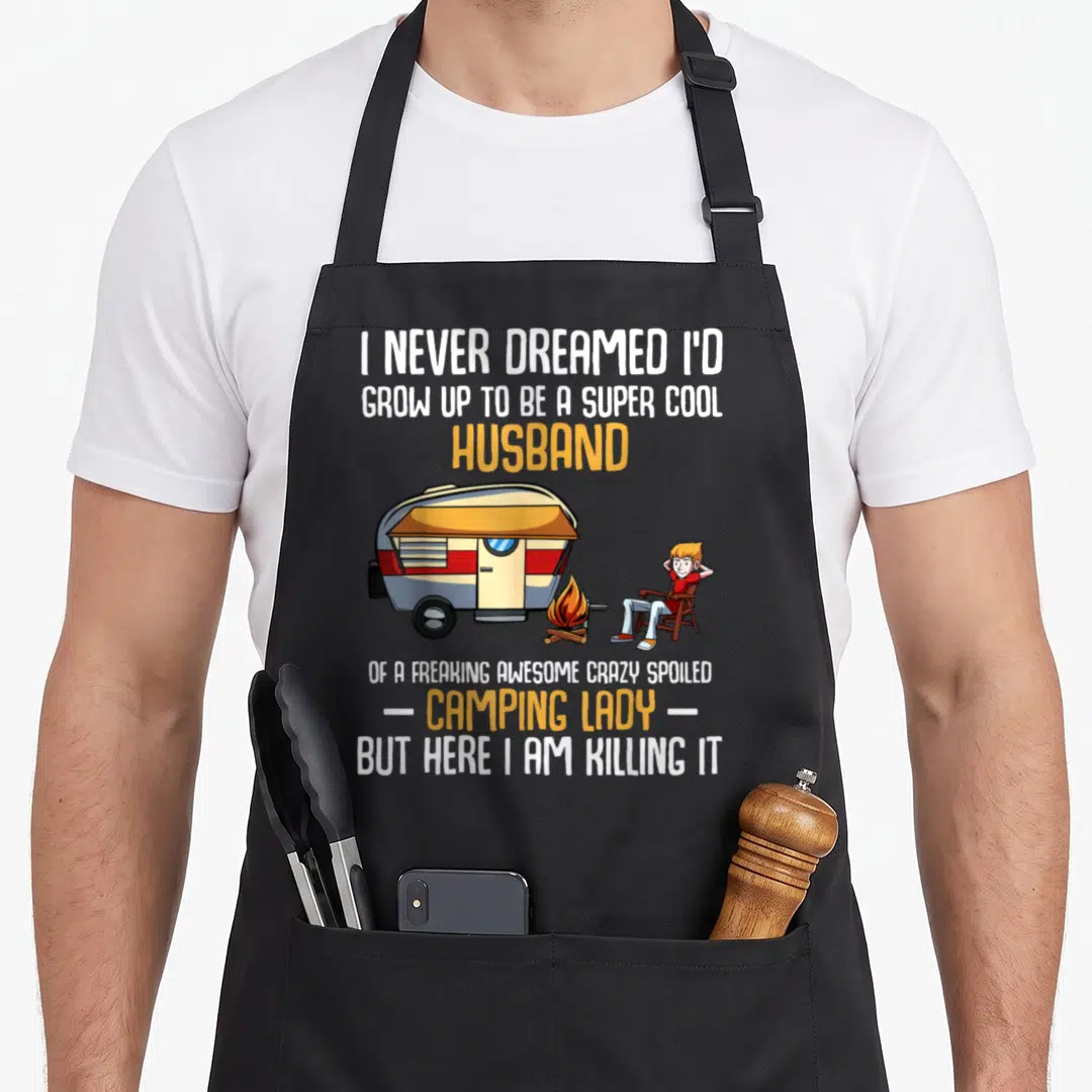 Dreamed husband apron - Cartoon man by camper trailer and campfire with sarcastic camping husband text — shown on a black cotton canvas apron against a white background