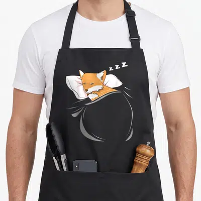 Sleeping fox aprons - Cartoon orange fox lying on white pillow under grey blanket with ZZZ text floating above head — shown on a black cotton canvas apron against a white background