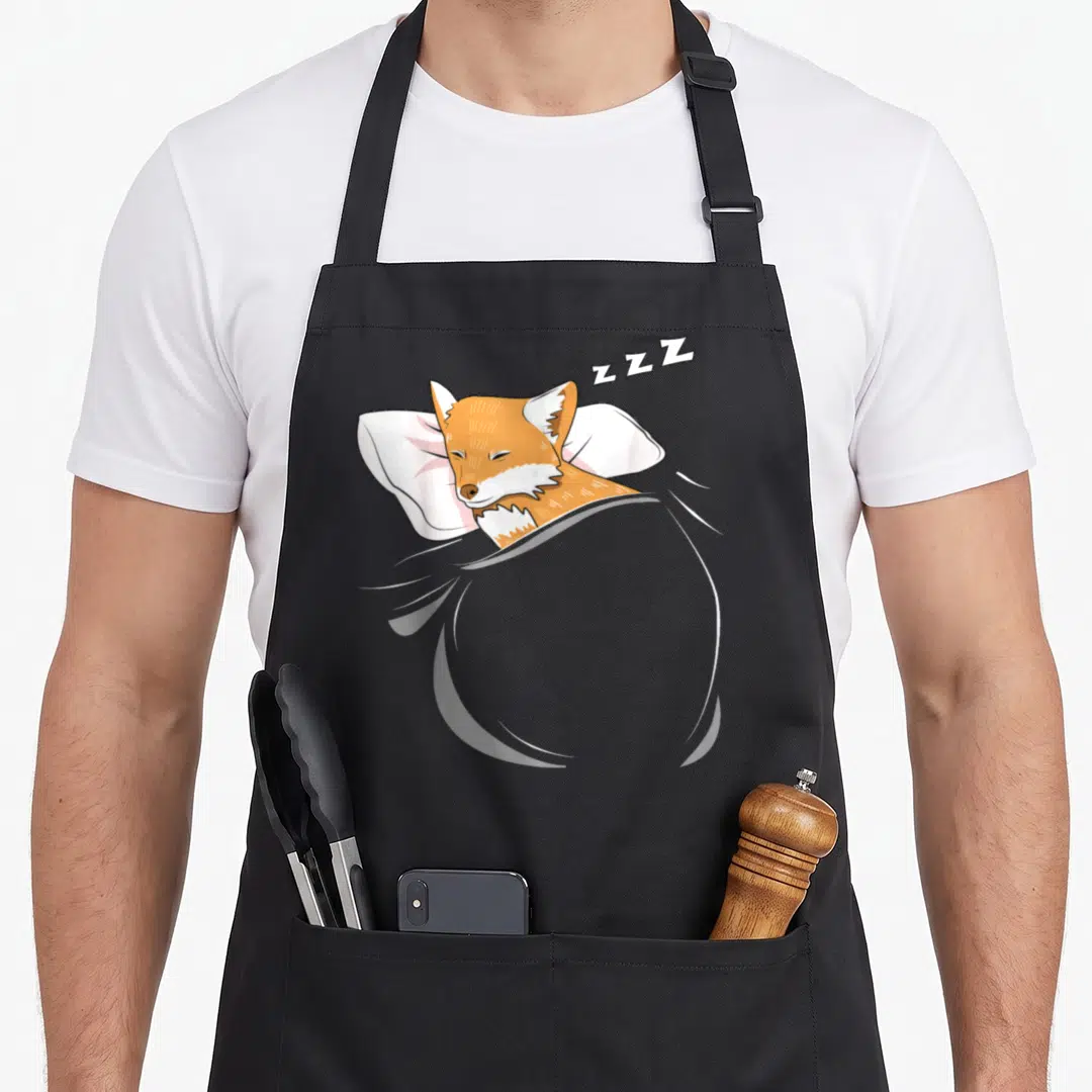 Sleeping fox aprons - Cartoon orange fox lying on white pillow under grey blanket with ZZZ text floating above head — shown on a black cotton canvas apron against a white background