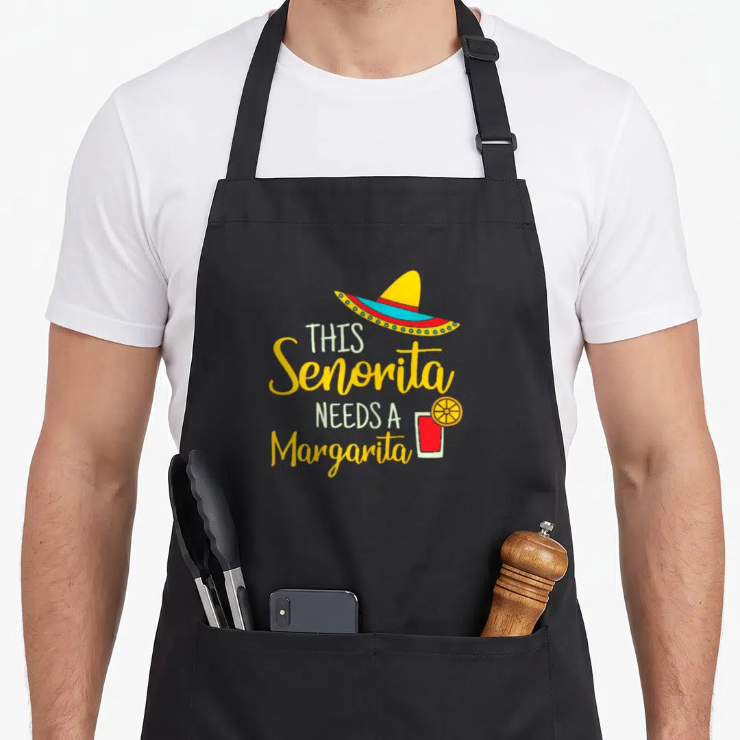 Senorita mexican apron - Stacked text THIS Senorita NEEDS A Margarita with sombrero and red drink with lime slice — shown on a black cotton canvas apron against a white background