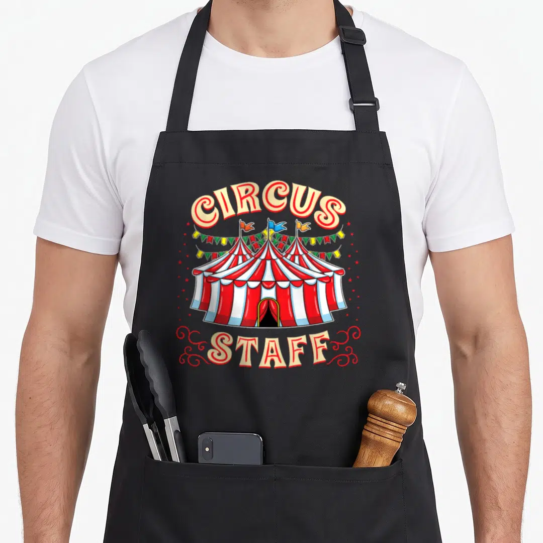 Funny circus staff aprons - Cartoon red and white striped circus tent with CIRCUS STAFF text and bunting — shown on a black cotton canvas apron against a white background