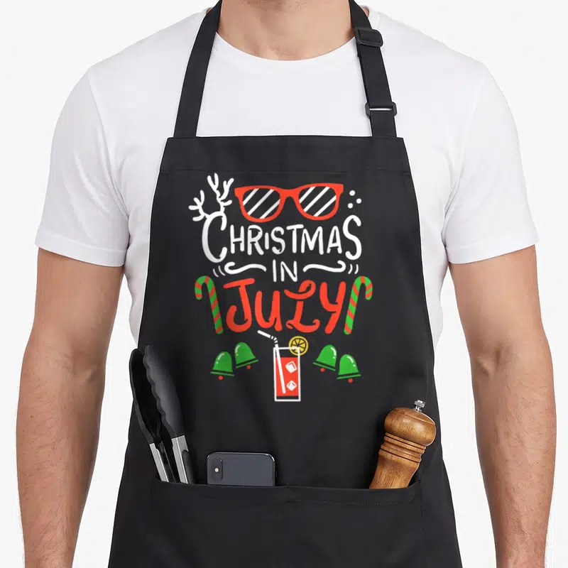 Summer christmas vacation apron - Cartoon text Christmas In July with red glasses, candy canes, drink, and four green — shown on a black cotton canvas apron against a white background