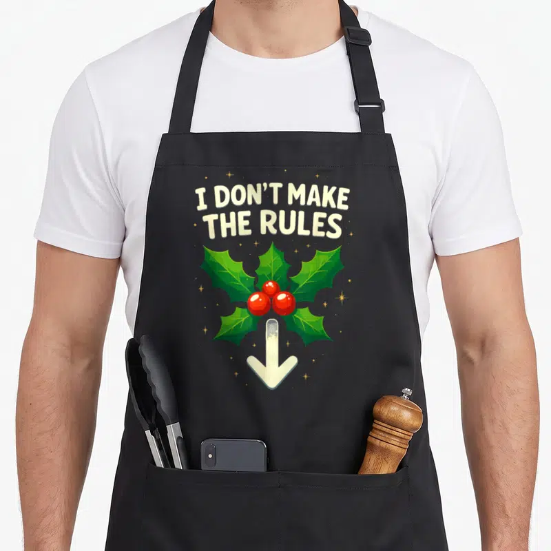 Inappropriate mistletoe christmas apron - I DON'T MAKE THE RULES text above holly leaves and downward arrow — shown on a black cotton canvas apron against a white background
