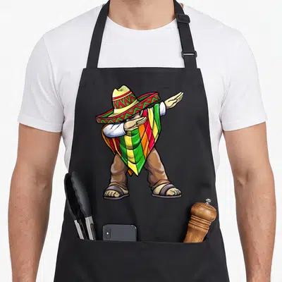 Mayo mexican apron - Cartoon man in yellow green red poncho and sombrero dabbing pose — shown on a black cotton canvas apron against a white background