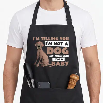 Dane mother's day aprons - Cartoon brown dog sitting beside stacked text reading I'm not a dog my mom said I'm a baby — shown on a black cotton canvas apron against a white background