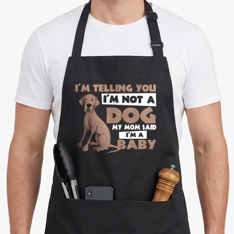 Dane mother's day aprons - Cartoon brown dog sitting beside stacked text reading I'm not a dog my mom said I'm a baby — shown on a black cotton canvas apron against a white background