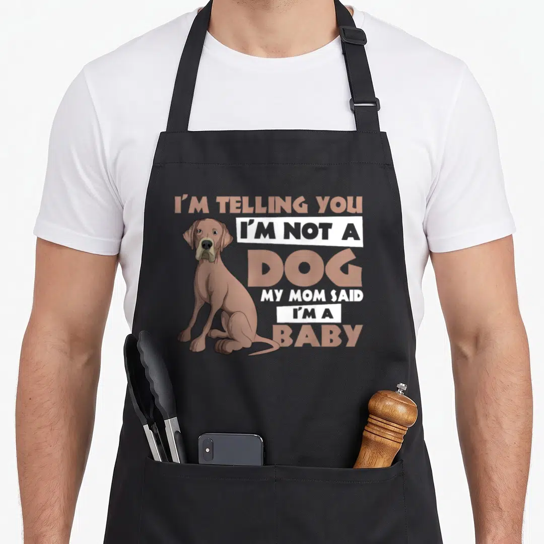 Dane mother's day aprons - Cartoon brown dog sitting beside stacked text reading I'm not a dog my mom said I'm a baby — shown on a black cotton canvas apron against a white background