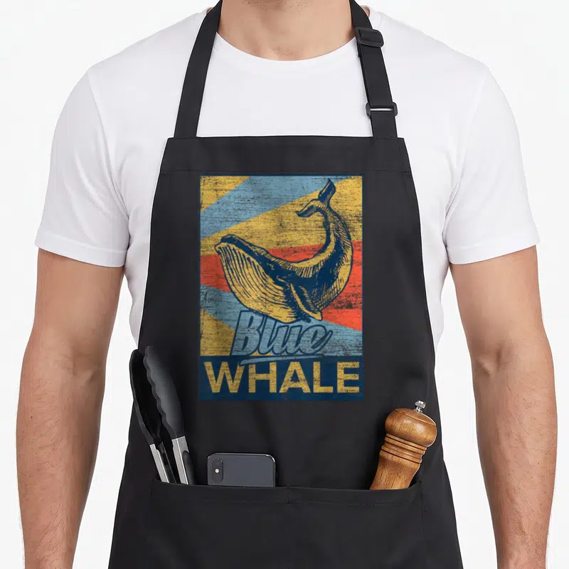 Blue whale animal apron - Retro yellow whale illustration over blue, yellow, and red rays with Blue WHALE text — shown on a black cotton canvas apron against a white background
