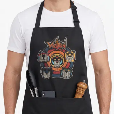 Voltron lion apron - VOLTRON logo above five colored robot heads in retro circle design — shown on a black cotton canvas apron against a white background