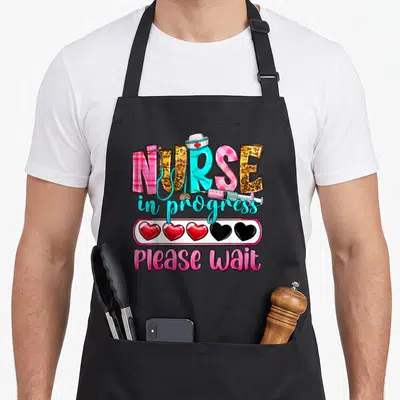Progress nurse apron - NURSE text with hat stethoscope syringe and heart loading bar in pink cyan yellow red cartoon — shown on a black cotton canvas apron against a white background