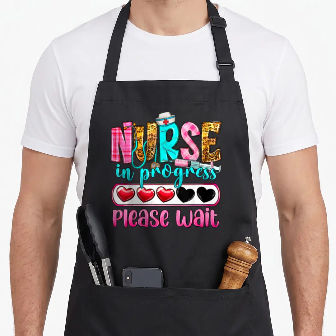 Progress nurse apron - NURSE text with hat stethoscope syringe and heart loading bar in pink cyan yellow red cartoon — shown on a black cotton canvas apron against a white background