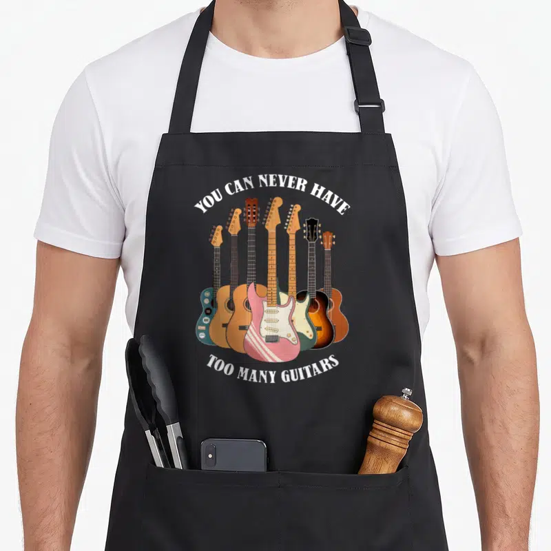 Acoustic crew apron - Seven cartoon guitars in pink blue brown yellow and white with arched white text — shown on a black cotton canvas apron against a white background