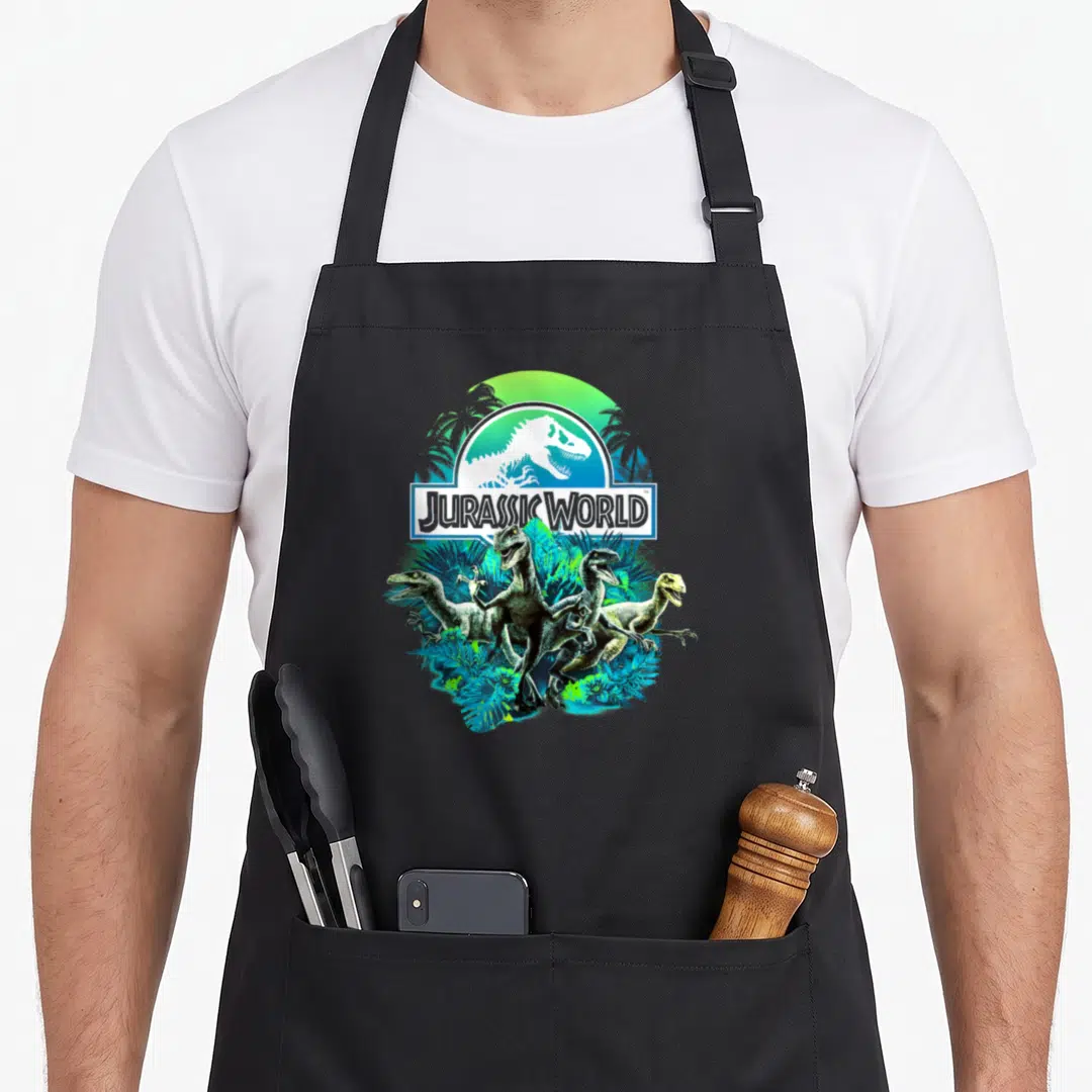 Jurassic velociraptor apron - Four raptors in blue and green jungle leaves below Jurassic World logo — shown on a black cotton canvas apron against a white background