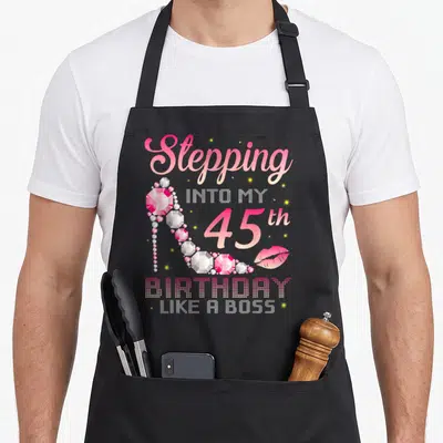 45th bbq boss apron - Pink diamond high heel shoe next to Stepping into my 45th birthday like a boss text — shown on a black cotton canvas apron against a white background