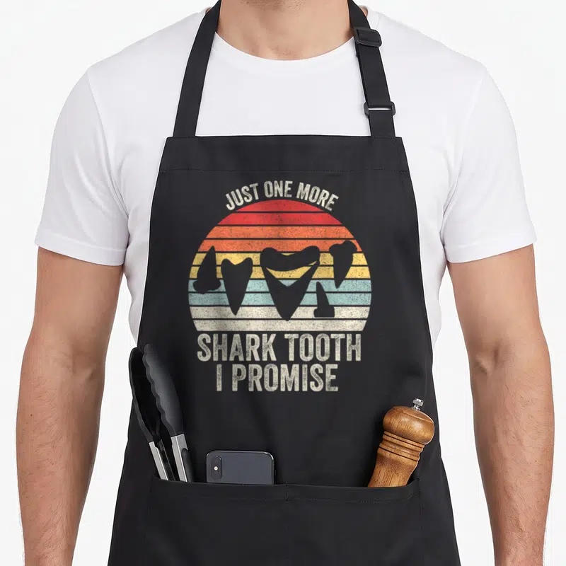 Tooth science apron - Retro striped sun with shark tooth silhouettes and text Just One More Shark Tooth I Promise — shown on a black cotton canvas apron against a white background