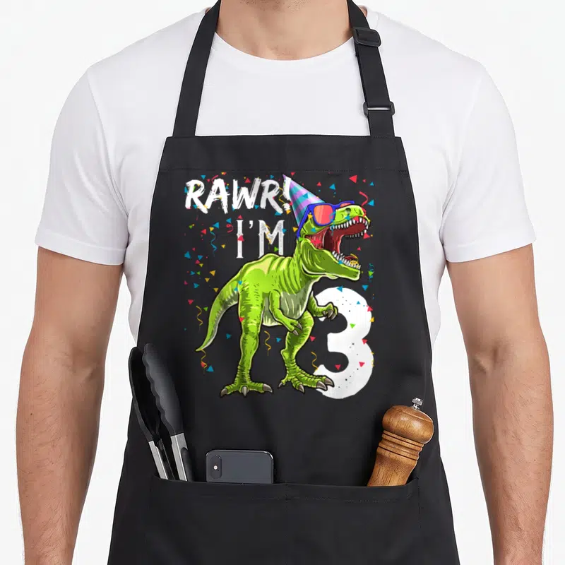 Rawr 3rd t rex apron - Green T-Rex with party hat sunglasses and number 3 surrounded by confetti — shown on a black cotton canvas apron against a white background