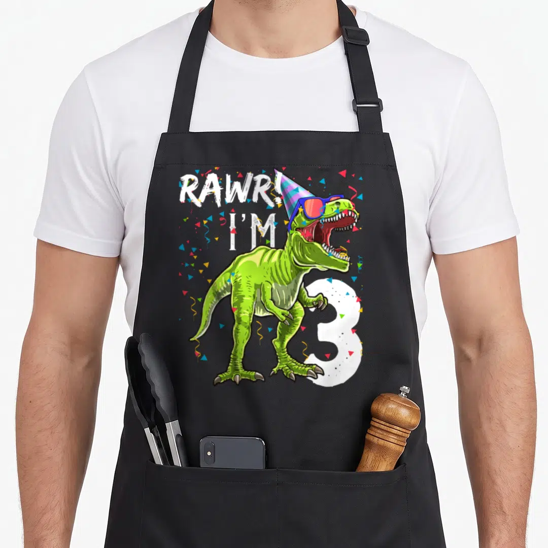 Rawr 3rd t rex apron - Green T-Rex with party hat sunglasses and number 3 surrounded by confetti — shown on a black cotton canvas apron against a white background