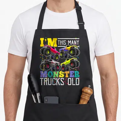 Many truck apron - Four monster trucks and confetti with I'M THIS MANY MONSTER TRUCKS OLD text — shown on a black cotton canvas apron against a white background